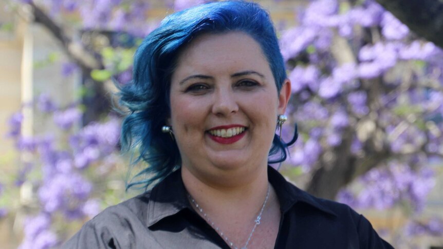 Headshot of child sex abuse campaigner and survivor Kirsty Pratt with blue hair against backdrop of jacarandas.