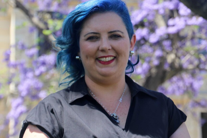 Headshot of child sex abuse campaigner and survivor Kirsty Pratt with blue hair against backdrop of jacarandas.