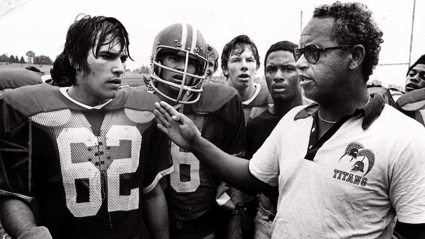 high school football players in uniform are coached by Herman Boone wearing a titans polo shirt and glasses
