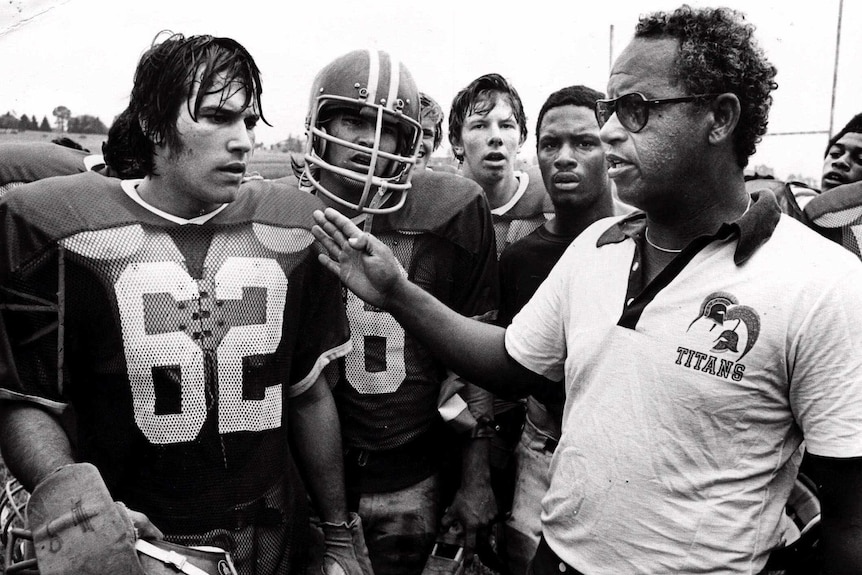 high school football players in uniform are coached by Herman Boone wearing a titans polo shirt and glasses