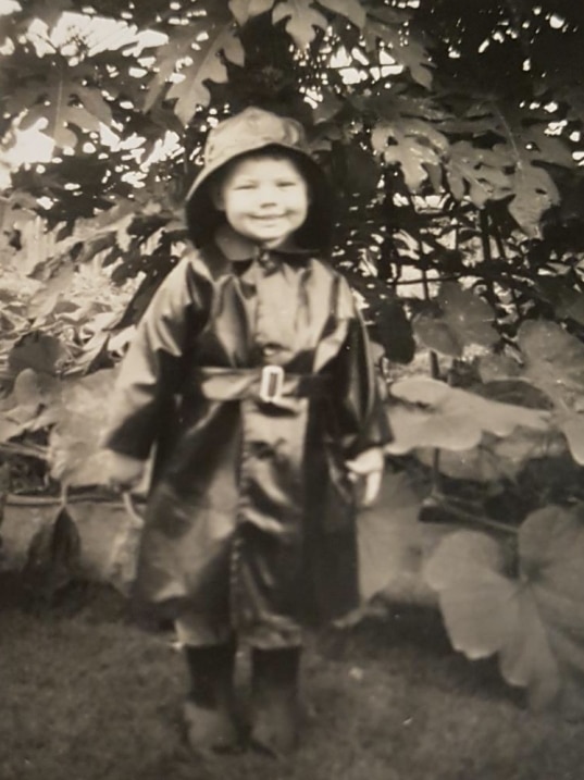A black and white image of a small boy standing in a rain coat in a garden.