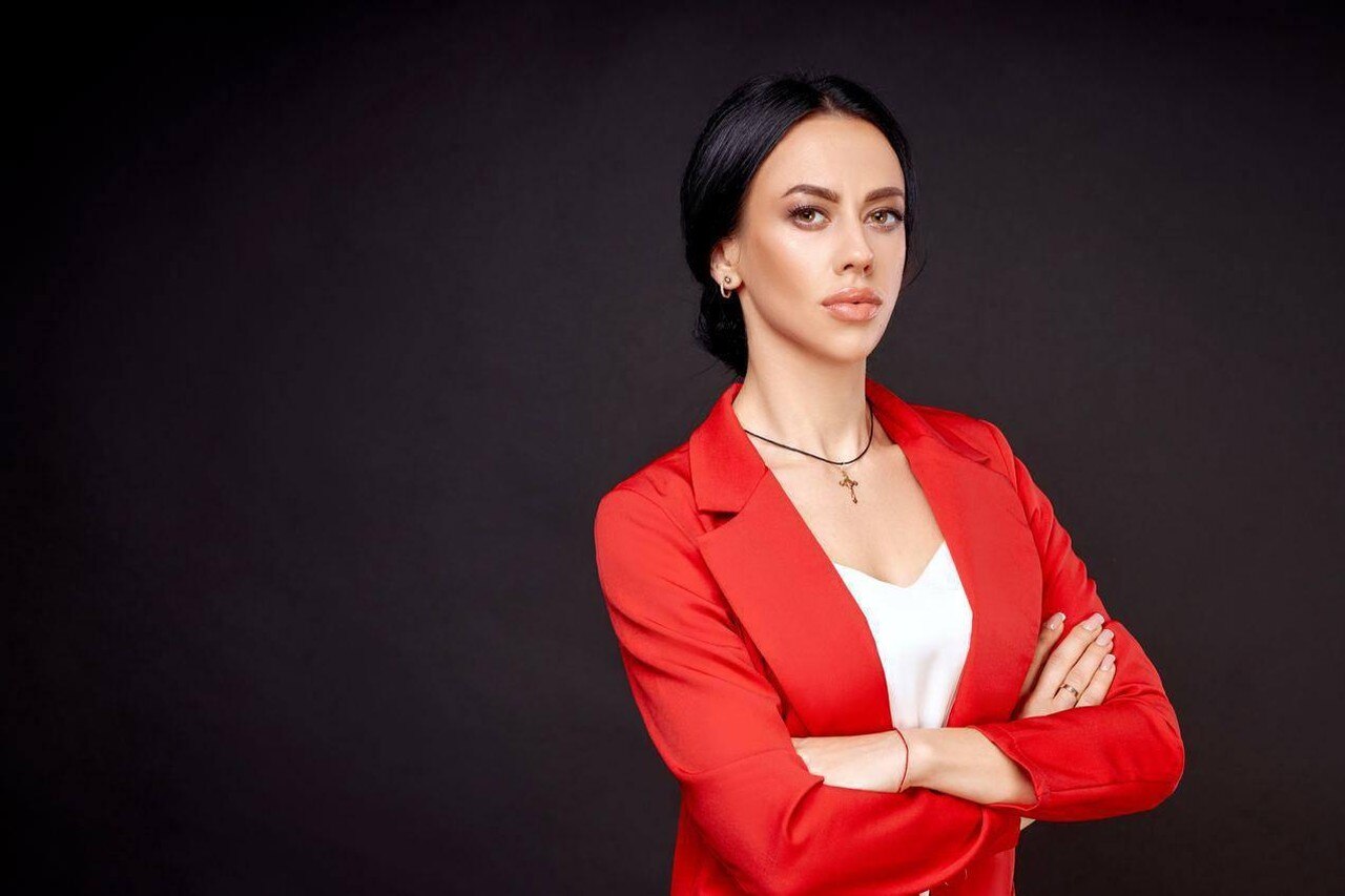 A young woman wearing red blazer over a white top, stands with arms folded across her chest