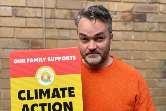 A man in an orange sweater holds a sign that says climate action now