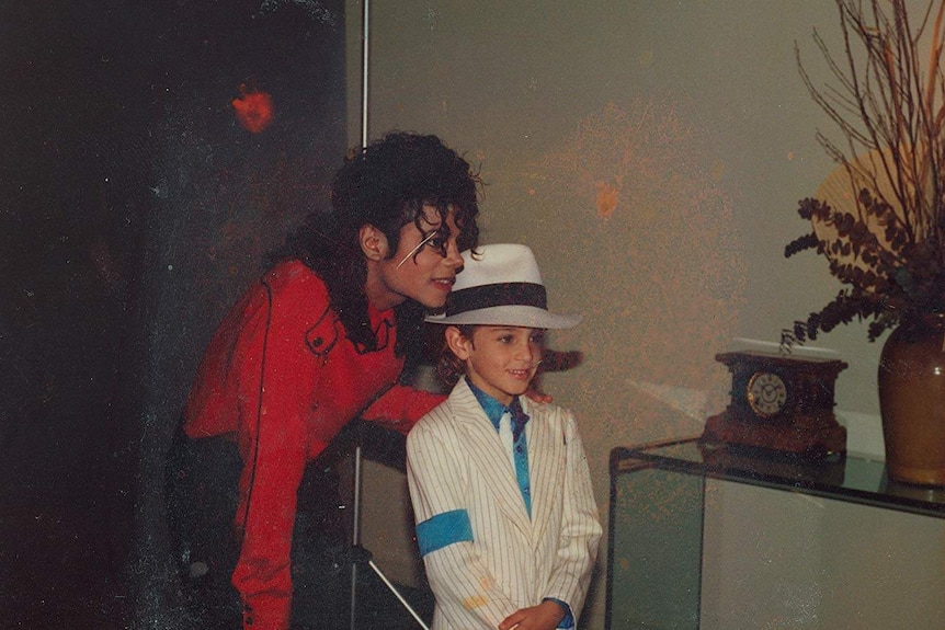 Michael Jackson, as an adult in a red jacket, leans down to put a hand on a child's shoulder. The child is in a pinstripe suit