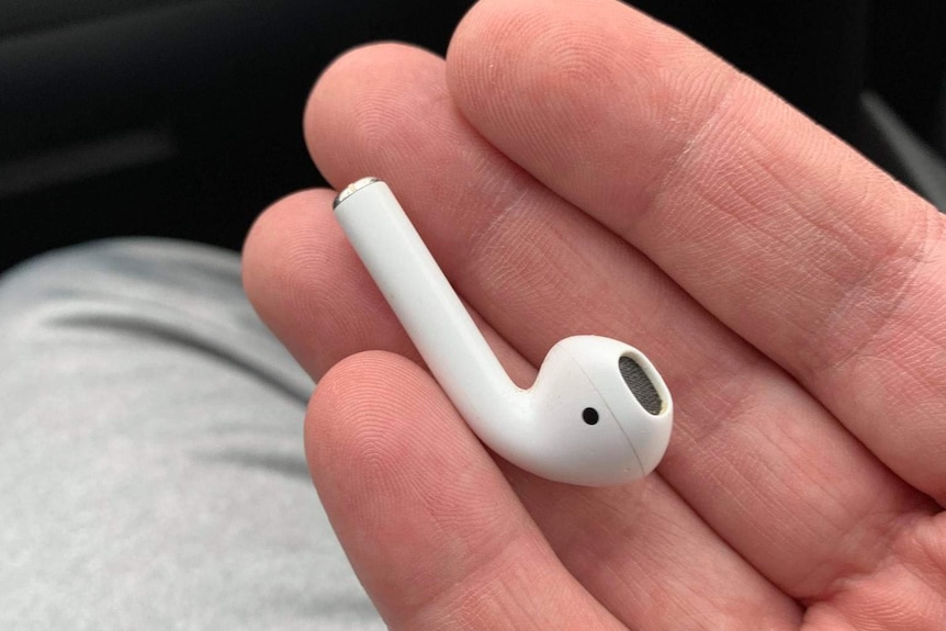 A single white wireless ear bud is held in a male hand in front of a black background.