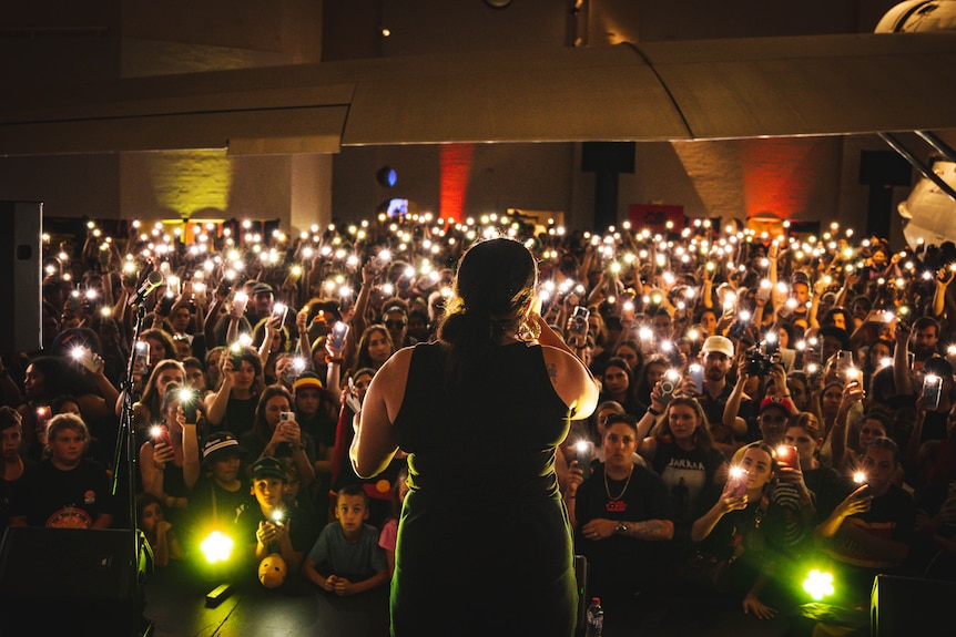 A woman stands on stage with her back to the camera singing, the audience are holding up their phone lights in a dark room
