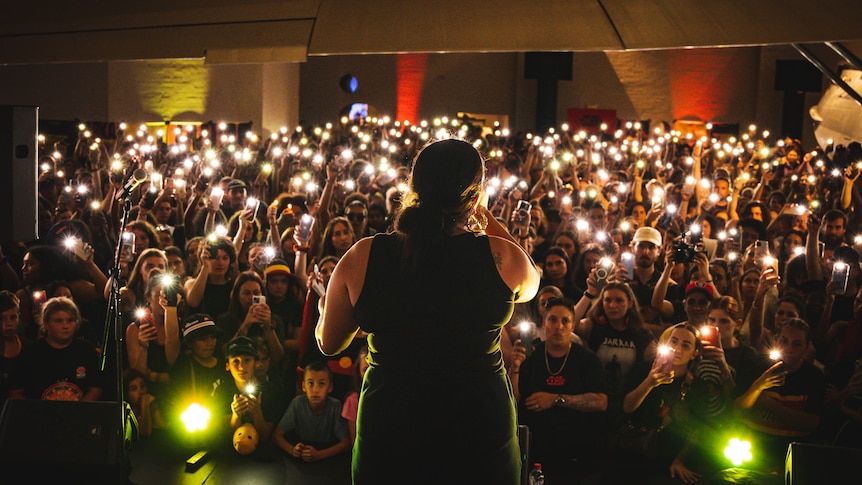 A woman stands on stage with her back to the camera singing, the audience are holding up their phone lights in a dark room