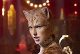 An fawn coloured CGI cat with woman's face stands in a dimly lit 1930s style cabaret-style music hall full of other cats.
