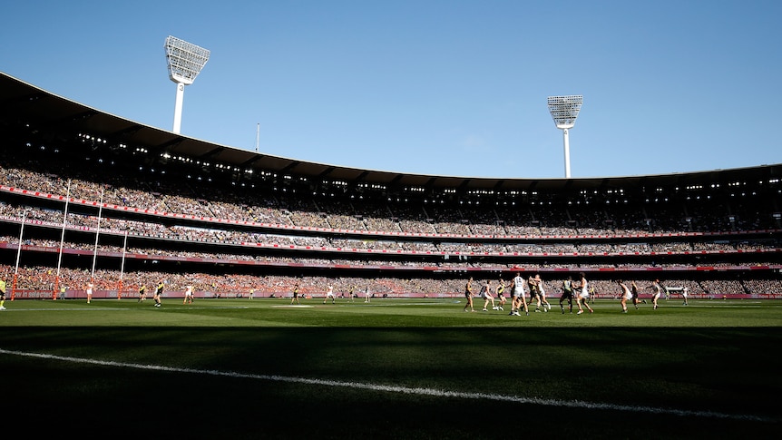The MCG stands filled on a clear, sunny day while a football game is played