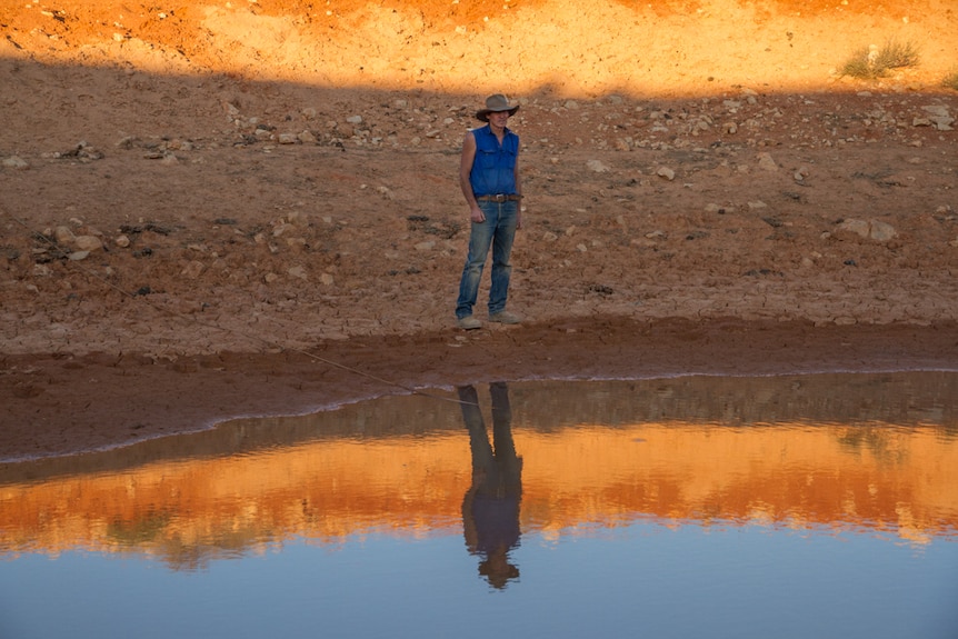 A man stands near a dam in the outback that has little water in it