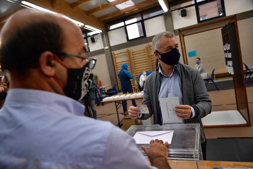 A grey-haired man with a face mask prepares to put a vote into a transparent box as a man watches.