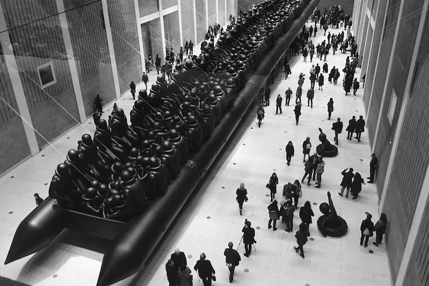 A large inflatable black raft about 70 metres long, filled with about 250 black vinyl figures, inside a large gallery hall