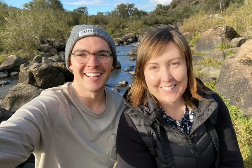 Young man wearing beanie, and woman, standing in front of creek.