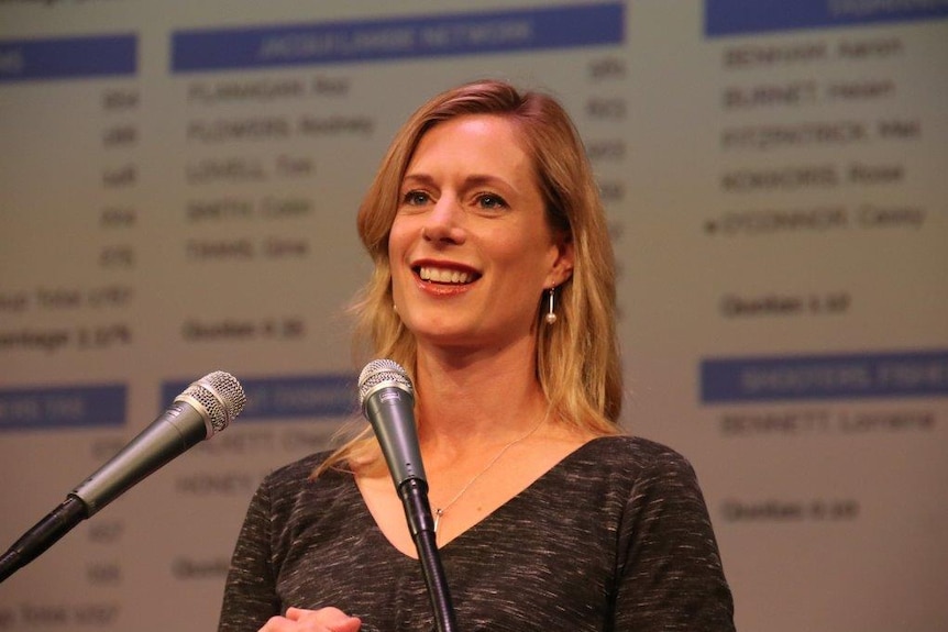 Rebecca White stands in front of a large screen displaying indistinguishable numbers on a tallyboard.