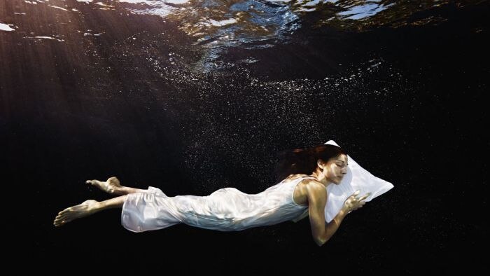 Woman in a white dress, eyes closed, head on pillow, underwater