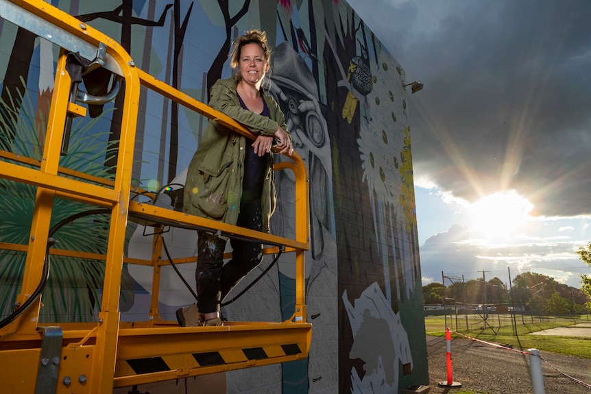 The sun sets on a woman in paint splashed black jeans and a khaki jacket, looks down at the camera from a scissor lift.