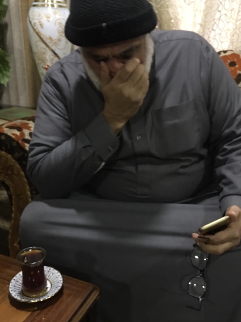 An imam sits in a floral armchair, with a cup of tea on the table in front. He is covering his face with one hand.