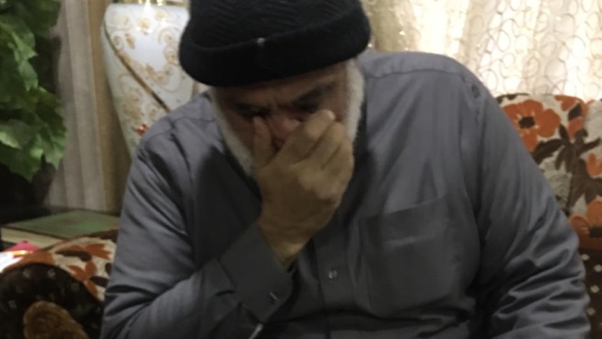 An imam sits in a floral armchair, with a cup of tea on the table in front. He is covering his face with one hand.