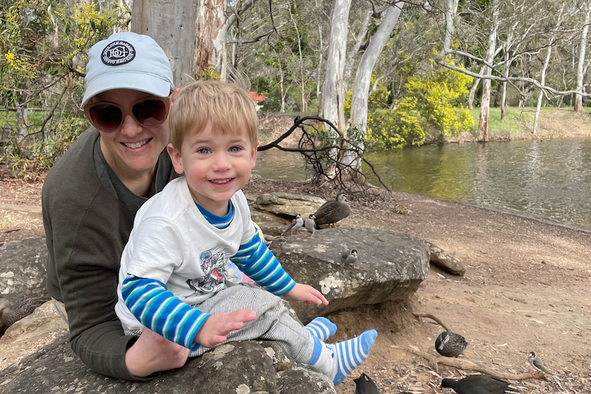 A woman in a cap and sunglasses next to a boy in white t-shirt and blue strips near water in a park