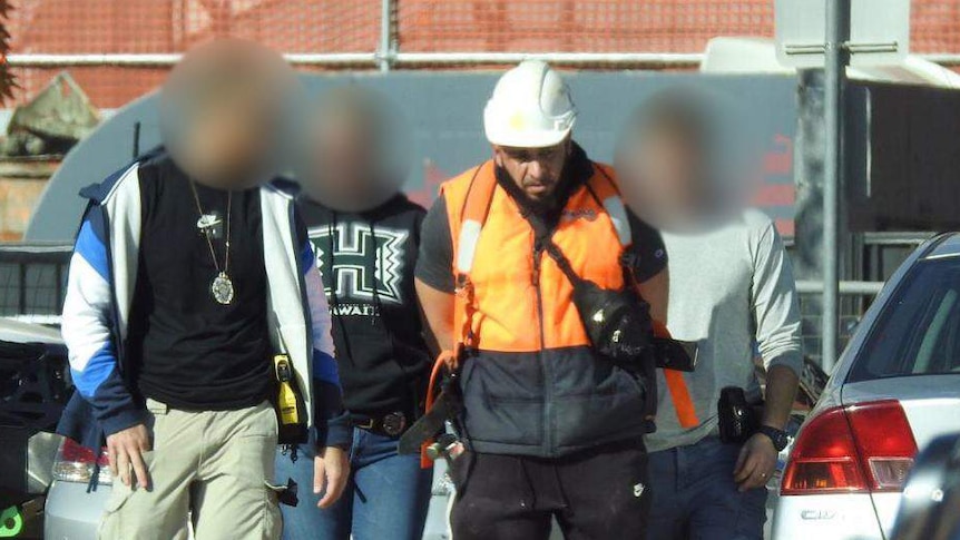 A man in high-visibility work gear is led in handcuffs by police officers.