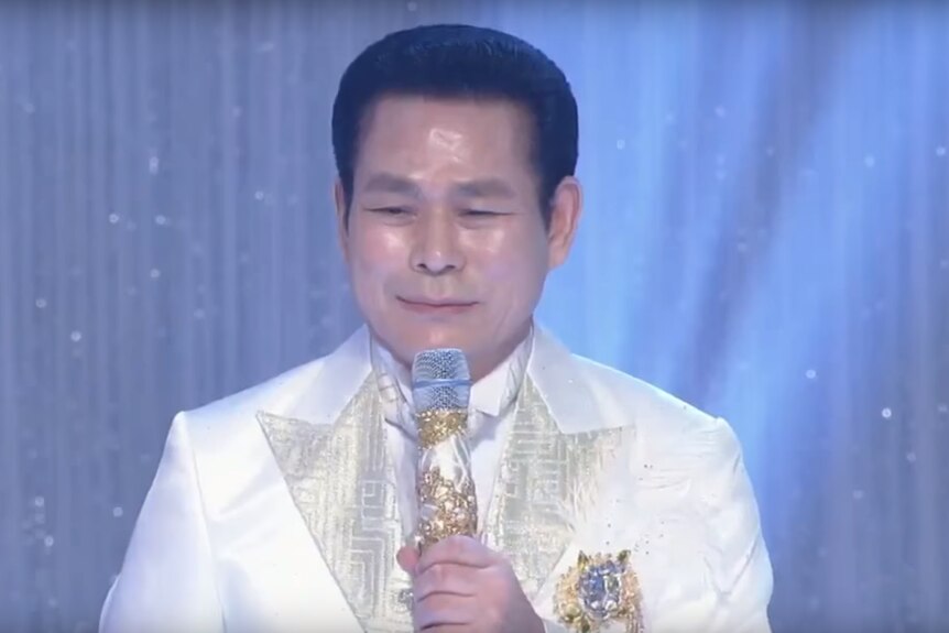 Manmin Central Church's founder Lee Jae-rock speaks to followers in a white suit holding an ornate microphone decorated in gold.
