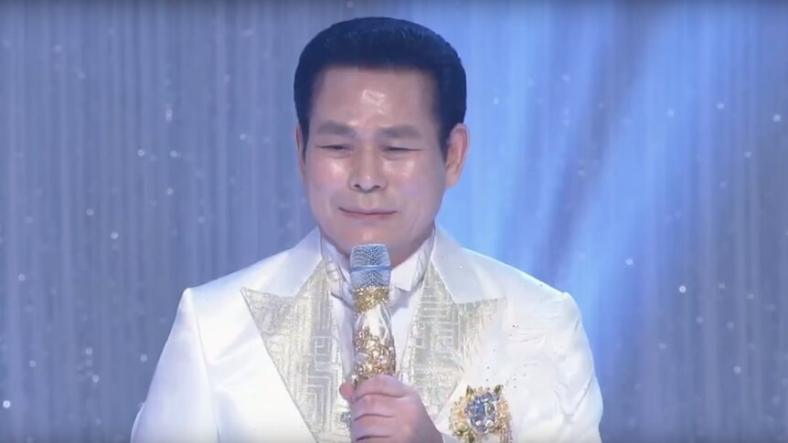 Manmin Central Church's founder Lee Jae-rock speaks to followers in a white suit holding an ornate microphone decorated in gold.