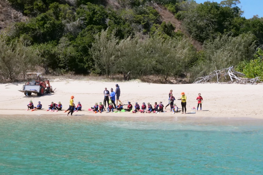 A group of people sit on the water's edge on the sand and put snorkel gear on.
