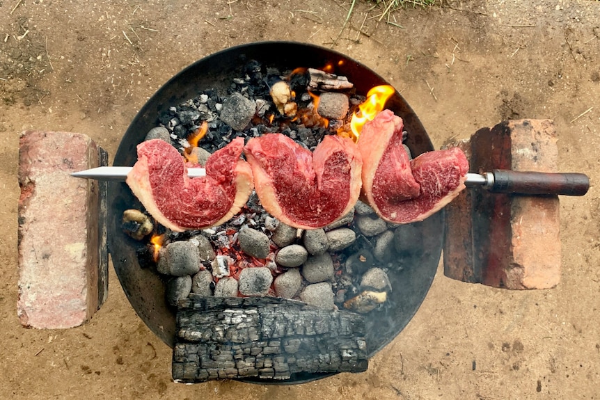 Cuts of meat skewered on a sword cook over a bucket of hot coals