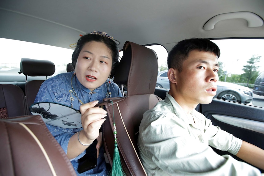 Inside a car, a woman in traditional Chinese leans over the centre console to speak to the driver.