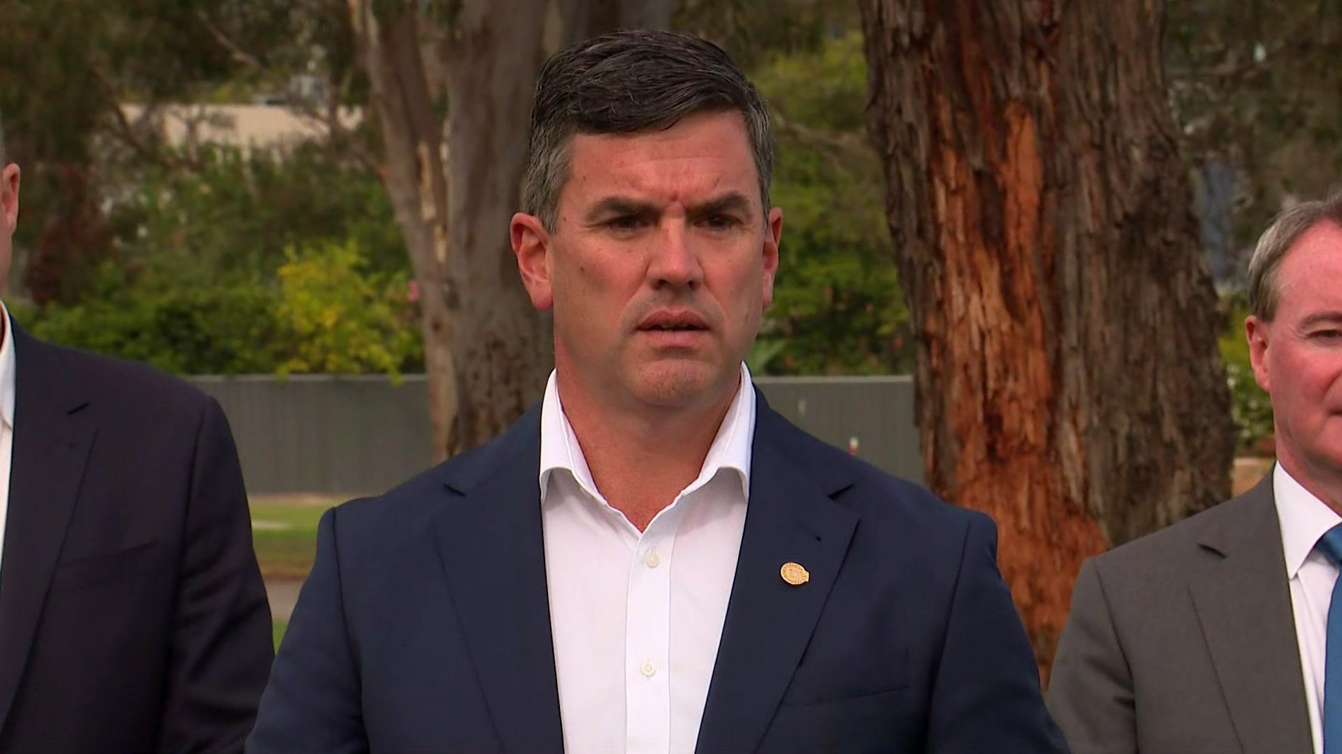 Um homem de cabelos escuros, vestindo uma jaqueta azul marinho e uma camisa de colarinho branco sem gravata olha para a câmera em um parque.