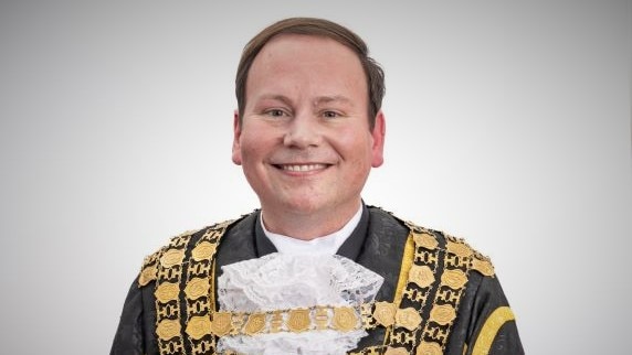 A portrait shot of a smiling man in gold and black mayoral robes.
