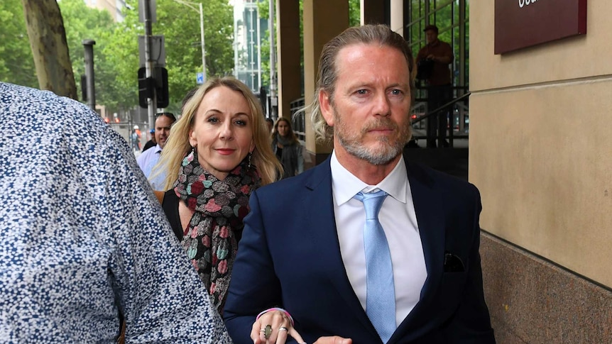 Craig McLachlan, who has a short beard and long hair, leaves court in a blue suit, next to his partner Vanessa Scammell.
