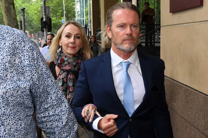 Craig McLachlan, who has a short beard and long hair, leaves court in a blue suit, next to his partner Vanessa Scammell.