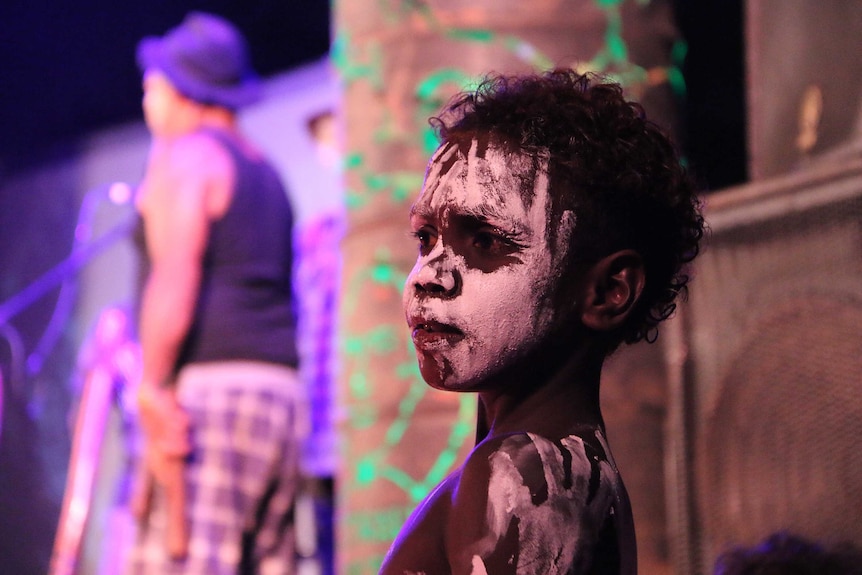 Young Indigenous boy with white face paint on looks away.