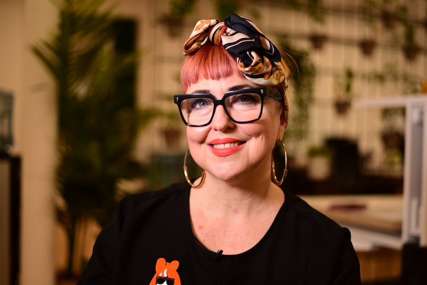 A woman with short pink hair, a silk bandana, black glasses, a black shirt and red lipstick smiles at the camera