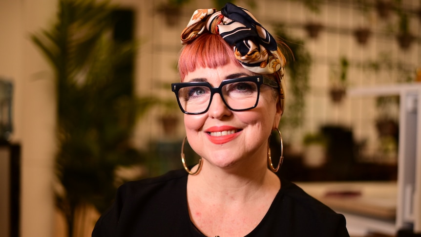 A woman with short pink hair, a silk bandana, black glasses, a black shirt and red lipstick smiles at the camera