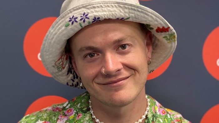 Queer artist Samuel Leighton Dore smiling in front of ABC Gold Coast sign wearing a white bucket hat and colourful shirt