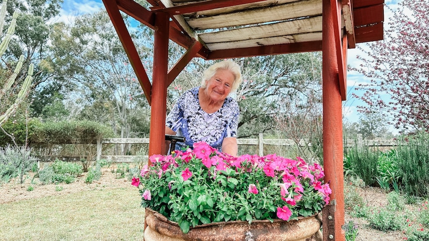 a woman stands behind a wishing well and a tub of purple flowers