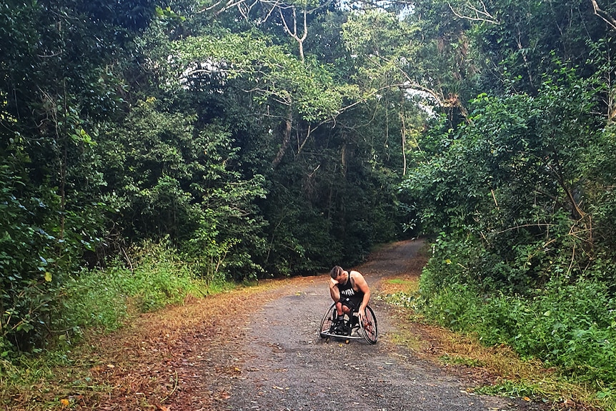 Man in wheel chair stopped with head in hand while climbing up steep gradient hill surrounded by bush.