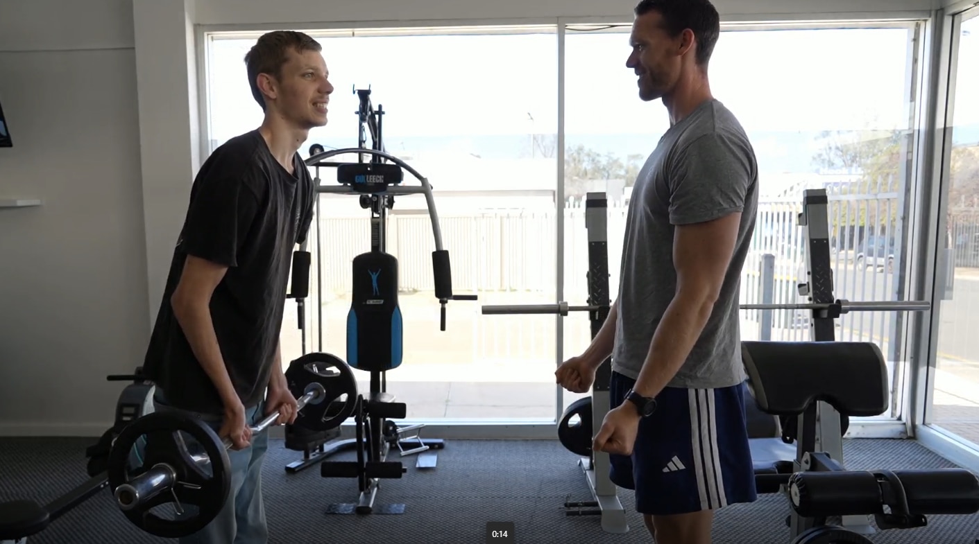 A young thin man lifting a barbell with weights on it, supported by another man opposite him.