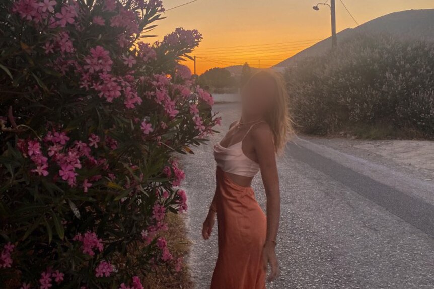 Girl in white crop top and long orange silk skirt stand and looks back at camera next to pink flower bush with the sunset