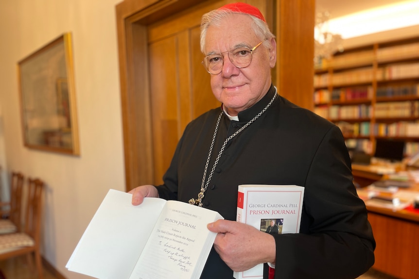 The Cardinal standing in front of a bookshelf holds open a book, showing a signed cover page.