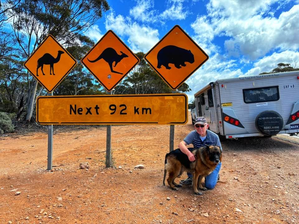 Lance crouching down with dog and caravan in the background on dirt road