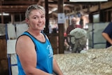A smiling blonde woman in a blue singlet stands in front of a fleece on a wool table
