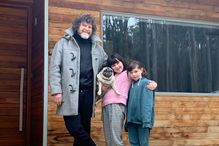 Man in grey jacket standing outside off grid home with two daughters and pug 