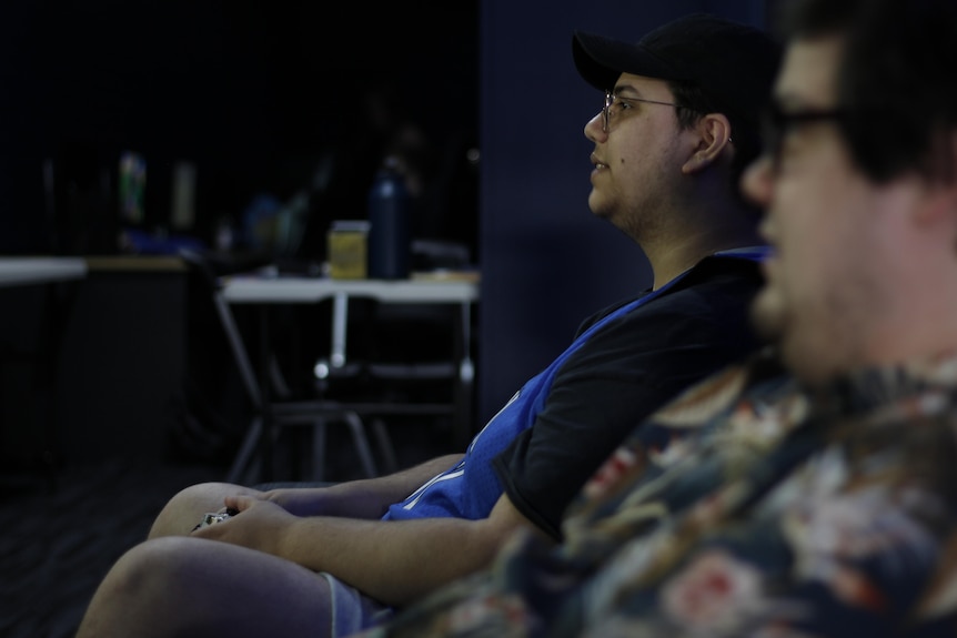 Two young men play video games together, with the camera focusing in on one wearing a black shirt and blue basketball jersey.