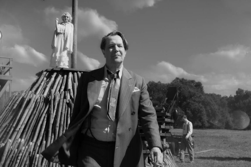Black and white film still showing Gary Oldman in 30s suit walking away from log pyre with blonde-haired actress on top.