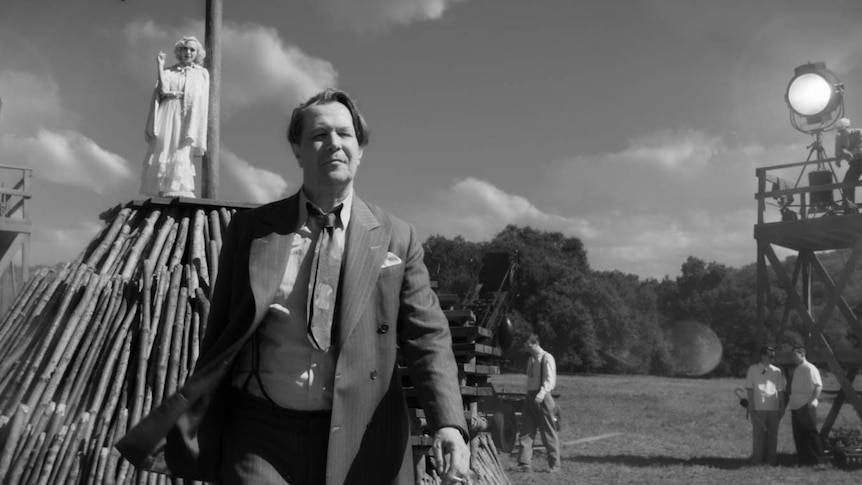 Black and white film still showing Gary Oldman in 30s suit walking away from log pyre with blonde-haired actress on top.