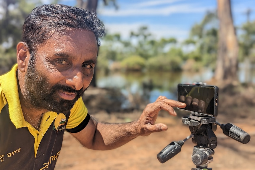 Close up of a Sikh Indian man with short hair and a short beard touching a mobile phone camera on a tripod by a river.