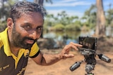 Close up of a Sikh Indian man with short hair and a short beard touching a mobile phone camera on a tripod by a river.